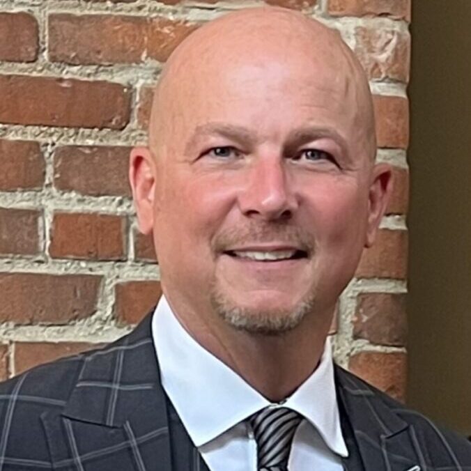 A bald man in a suit and tie standing in front of a brick wall.